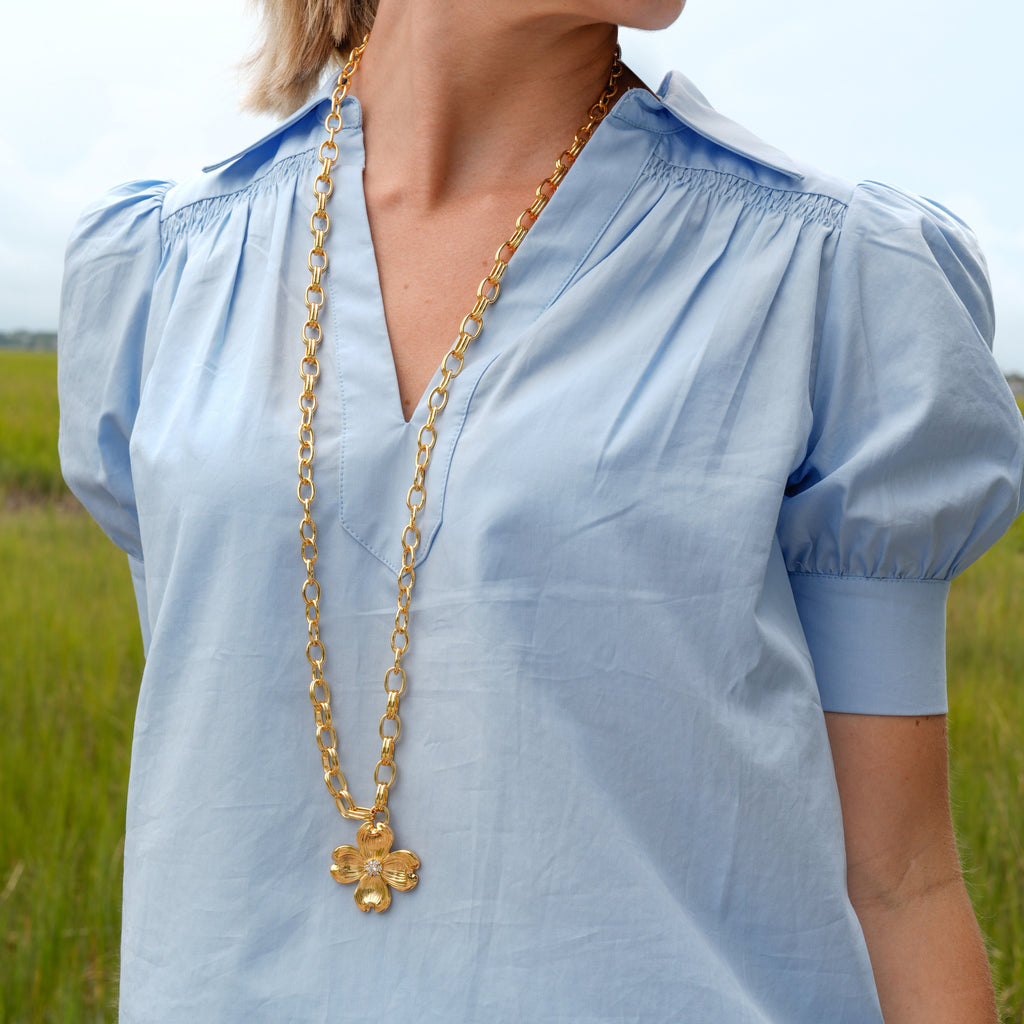 A woman wearing a light blue short-sleeve blouse with a smocked V-neck and puff sleeves, styled with a gold chain necklace, photographed outdoors in a grassy field.