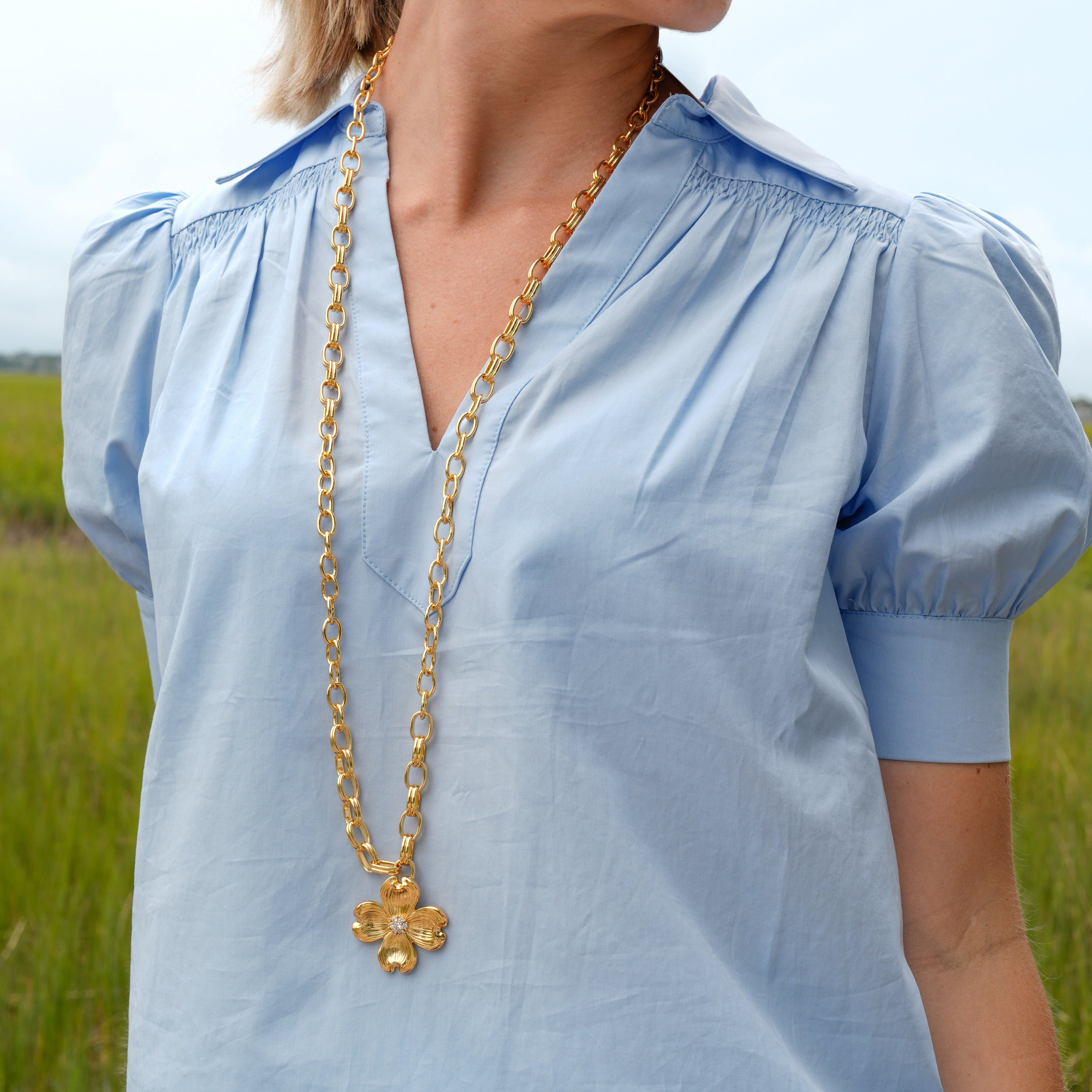 A woman wearing a light blue short-sleeve blouse with a smocked V-neck and puff sleeves, styled with a gold chain necklace, photographed outdoors in a grassy field.