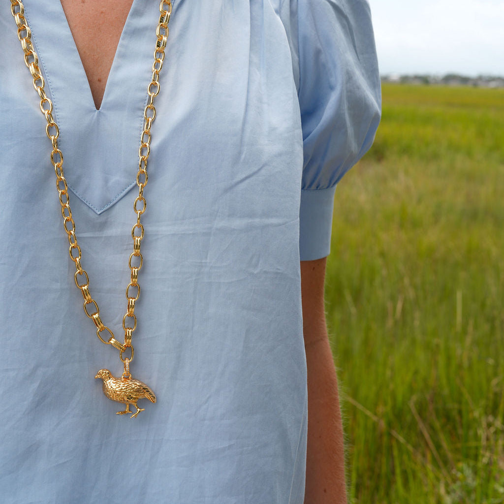 A close-up of a person wearing a light blue blouse and a long gold chain necklace with a small bird pendant, standing outdoors in a grassy field.
