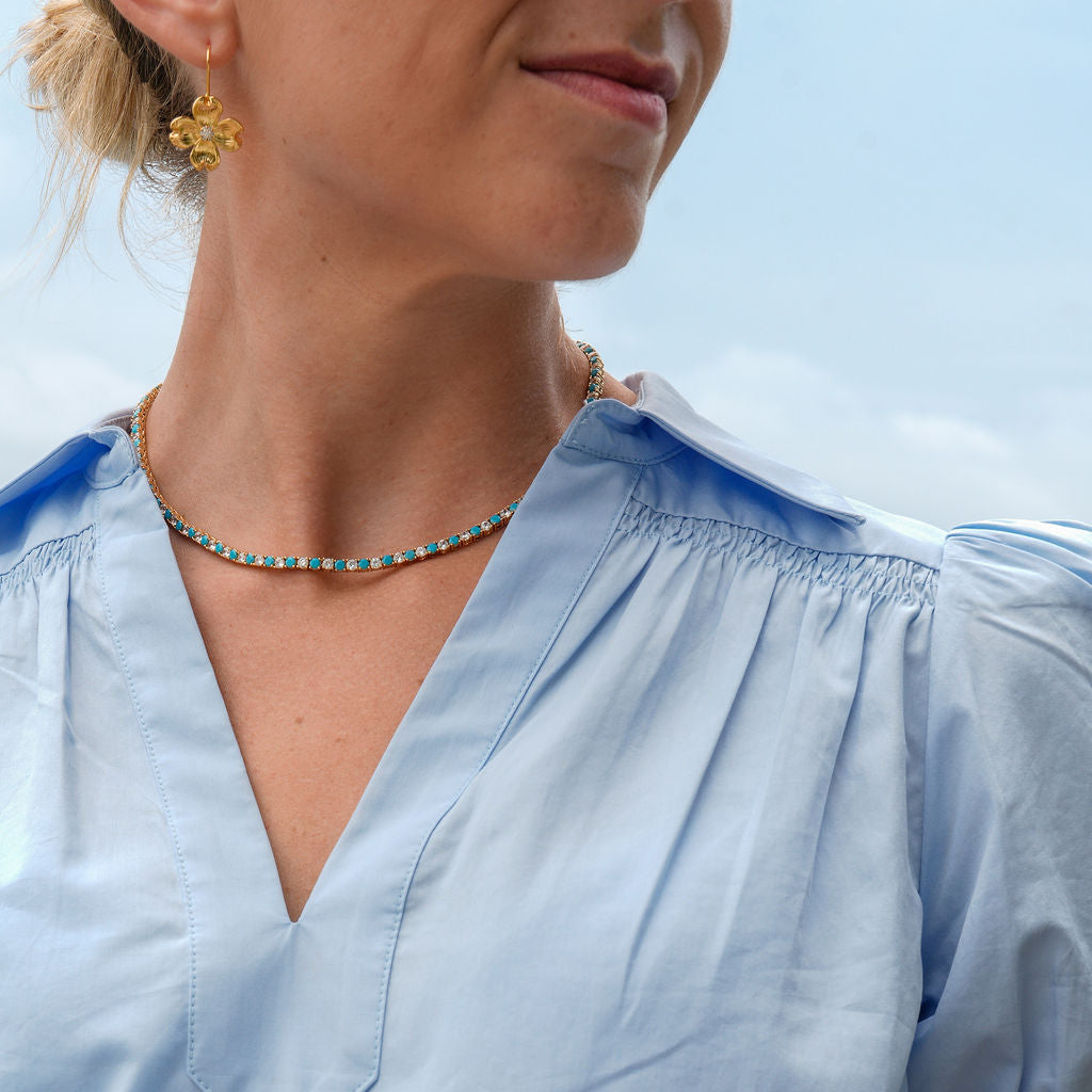 A close-up of a woman wearing a light blue short-sleeve blouse with a smocked V-neckline, paired with a beaded necklace and gold earrings against a pale blue sky background.