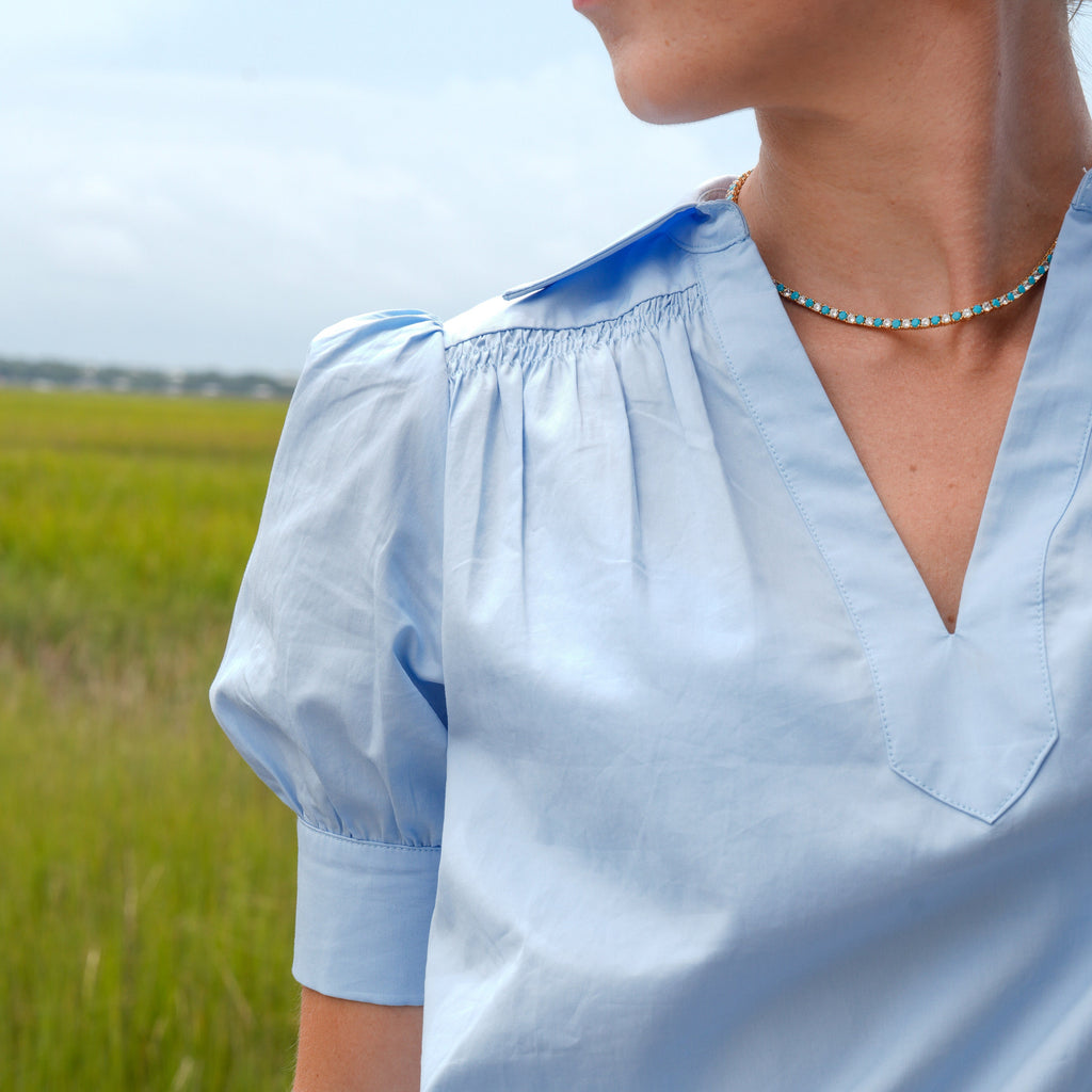 A close-up side profile of a woman wearing a light blue short-sleeve blouse with a smocked neckline and V-neckline, set against a blurred green field background.