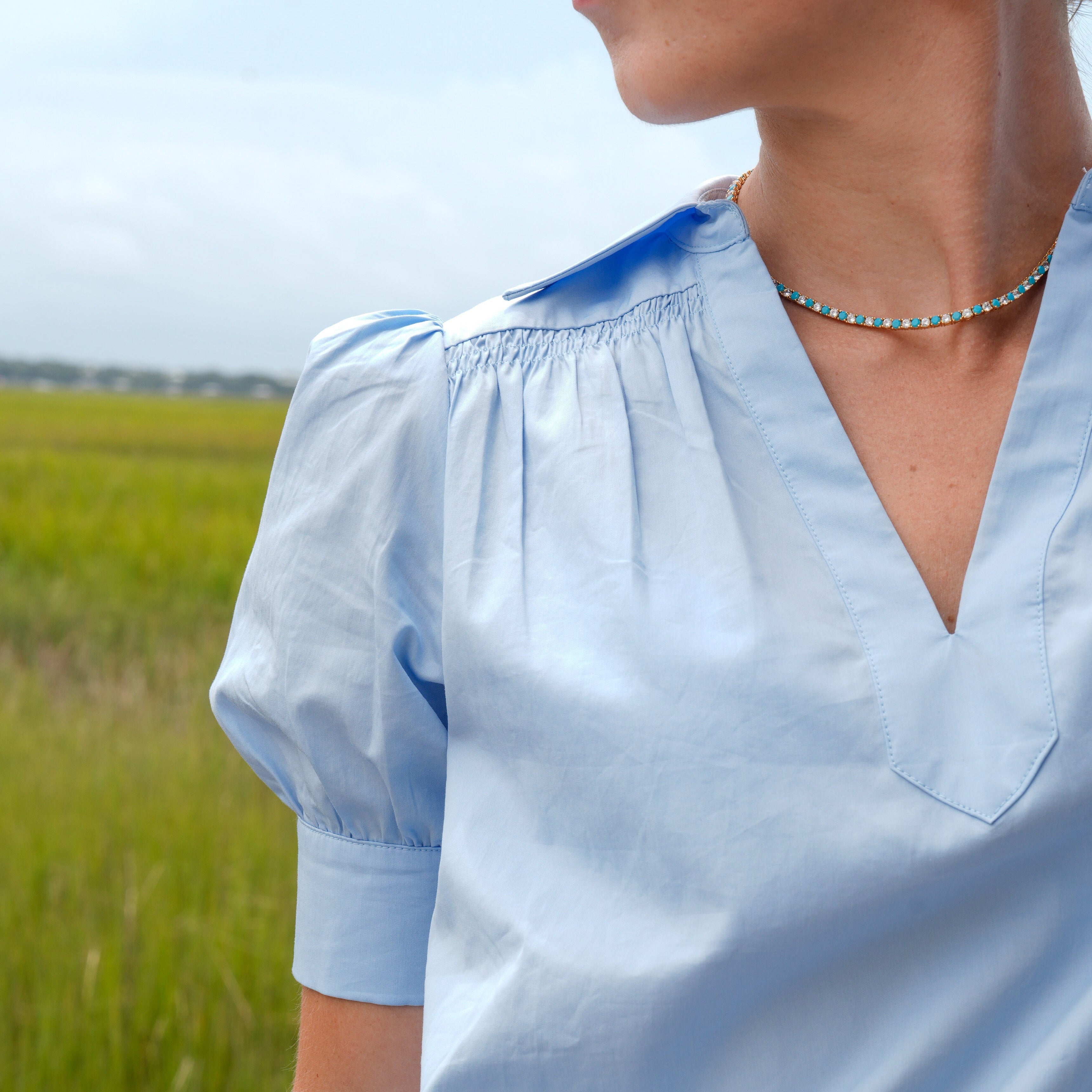 A close-up side profile of a woman wearing a light blue short-sleeve blouse with a smocked neckline and V-neckline, set against a blurred green field background.