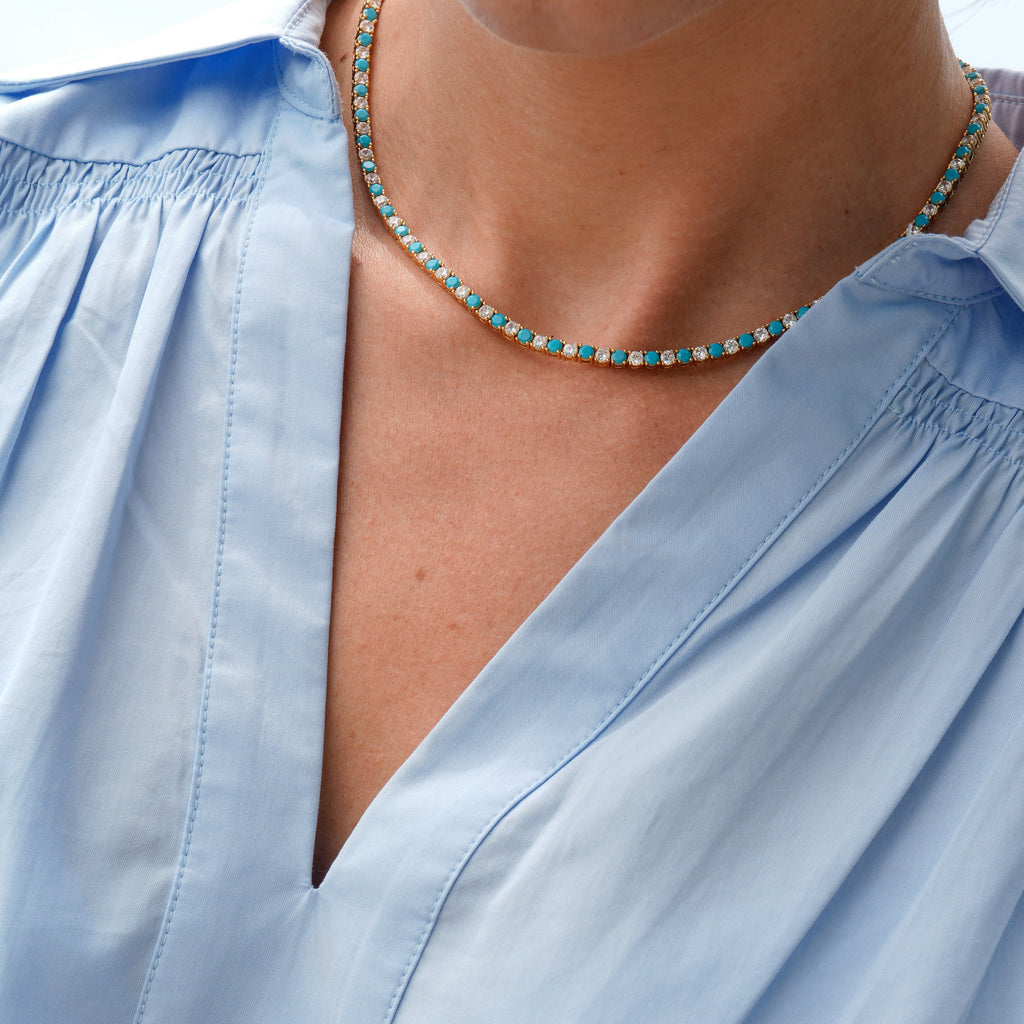 A close-up of a person wearing a delicate beaded necklace with turquoise blue and white beads, paired with a light blue blouse.