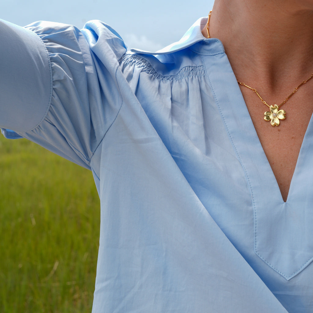 A close-up of a person outdoors wearing a light blue blouse with a gathered neckline and puff sleeves, paired with a gold flower pendant necklace, against a blurred green field background.