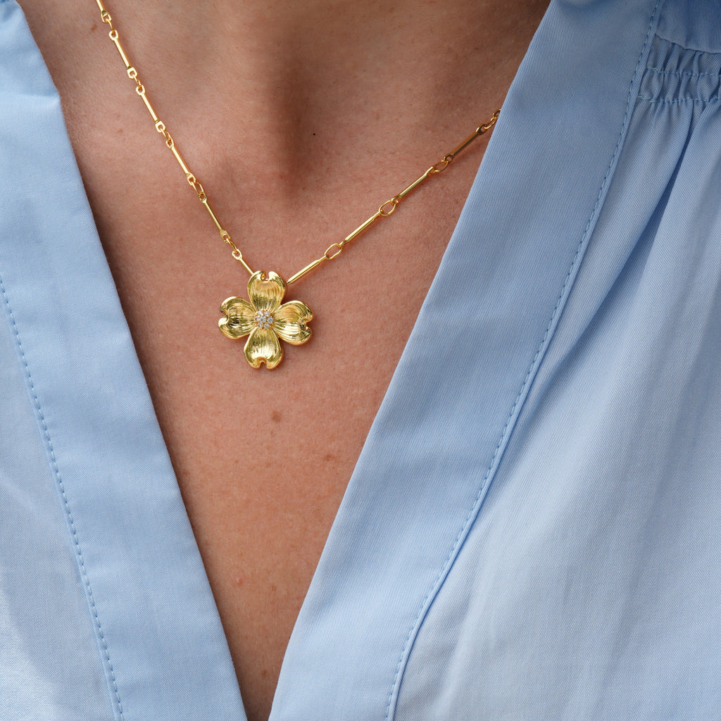 A close-up of a person wearing a gold flower-shaped pendant necklace with a clear faceted center, styled with a light blue V-neck blouse.