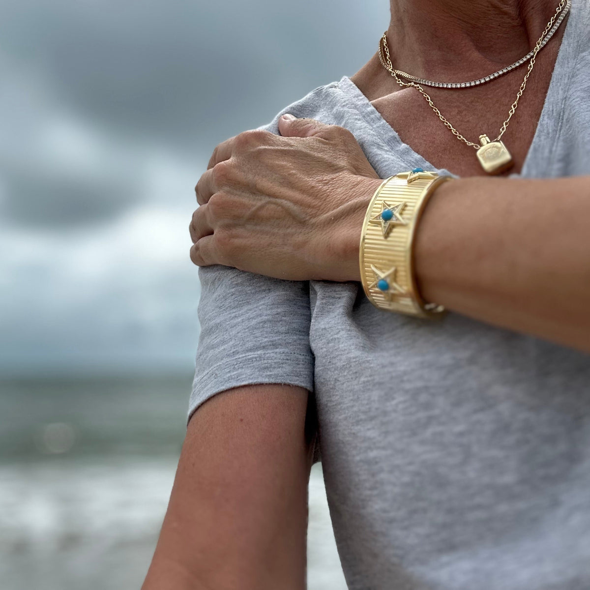 A close-up lifestyle shot of a person wearing a wide, textured gold cuff bracelet with blue star accents, layered with delicate gold necklaces, set against a blurred beach and cloudy sky background.