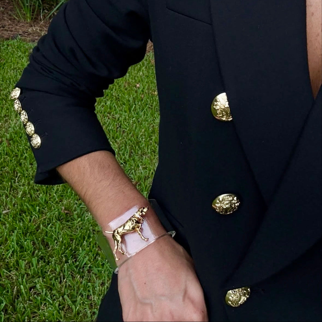 Woman standing with green grass in the background wearing a clear bracelet with a golden dog charm 