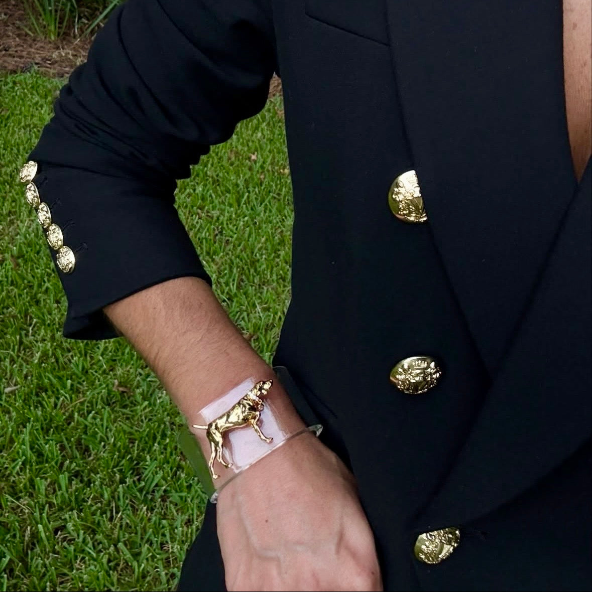 Woman standing with green grass in the background wearing a clear bracelet with a golden dog charm 