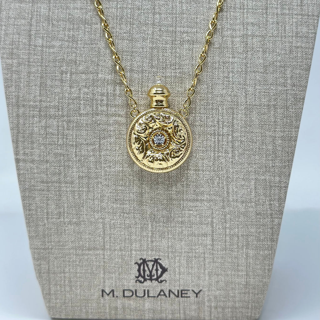 A gold pendant necklace displayed on a textured beige jewelry bust. The pendant is circular with ornate detailing and a small central clear gemstone.