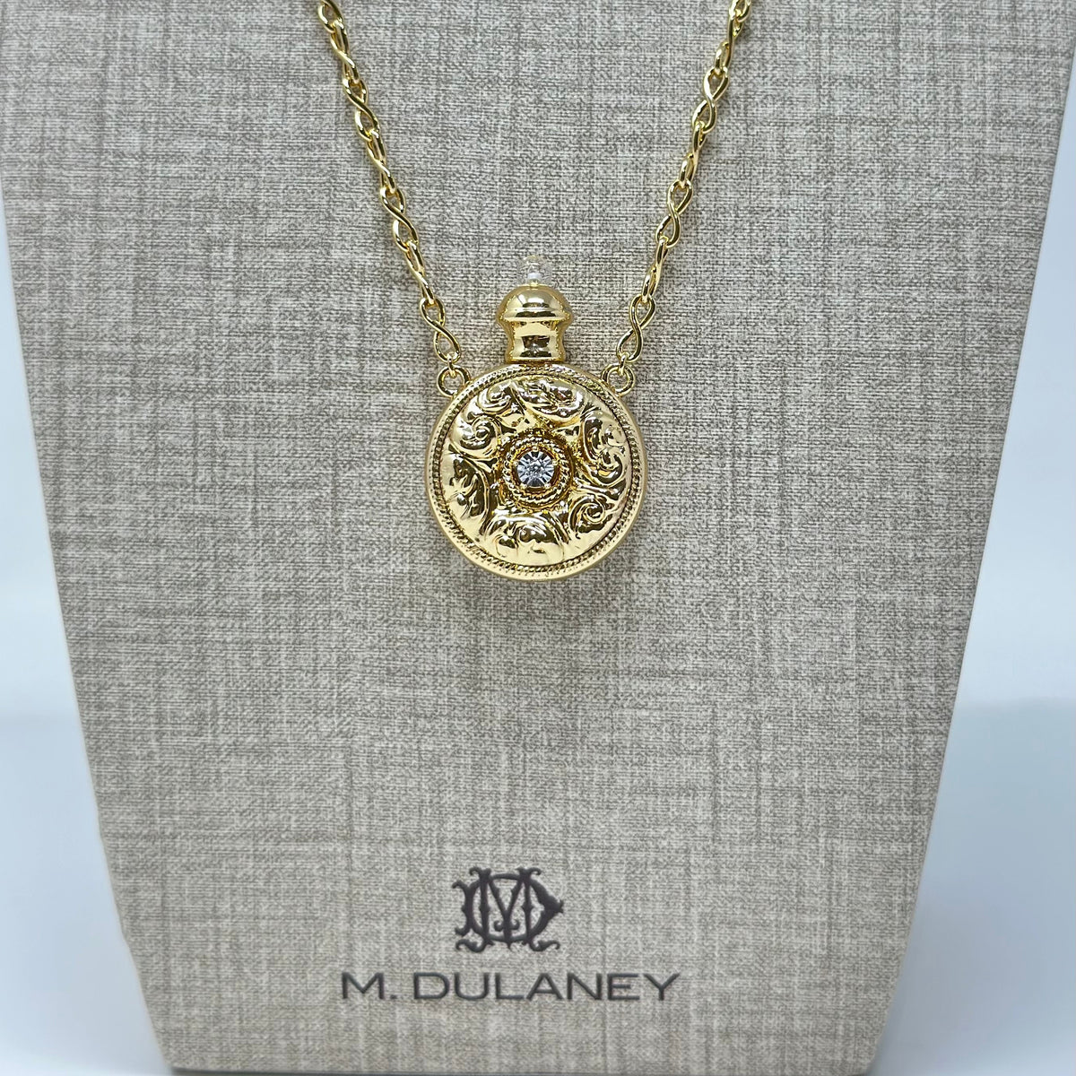 A gold pendant necklace displayed on a textured beige jewelry bust. The pendant is circular with ornate detailing and a small central clear gemstone.