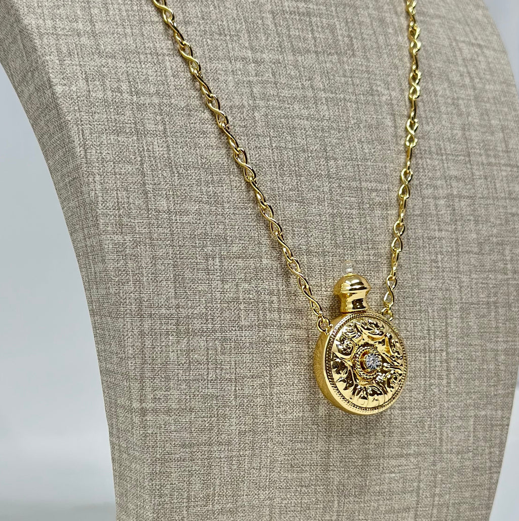 A gold locket necklace displayed on a beige fabric jewelry bust. The oval pendant features an intricate floral design with a small clear central stone.