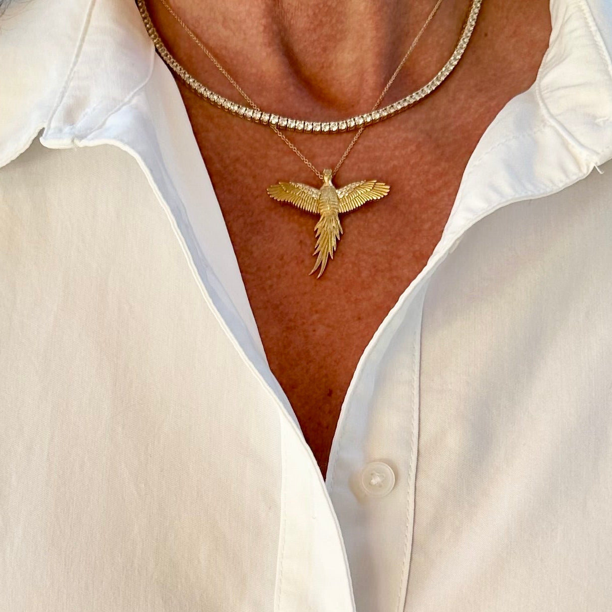 A close-up of a person wearing a white button-up shirt and layered necklaces, featuring a gold bird pendant on a beaded chain.