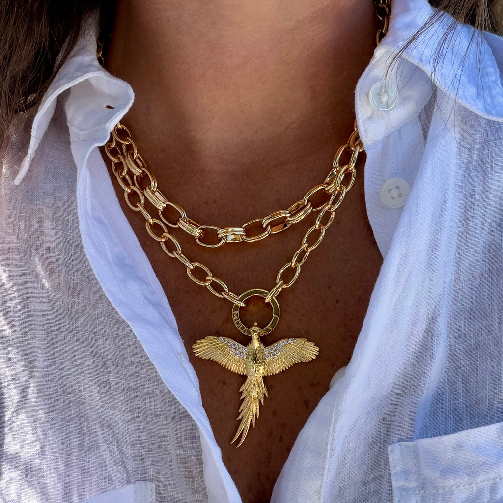 A close-up of a person wearing a layered gold necklace set, featuring a double-strand chain with a polished gold bird pendant, styled with a white linen button-up shirt.