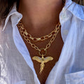 A close-up of a person wearing a layered gold necklace set, featuring a double-strand chain with a polished gold bird pendant, styled with a white linen button-up shirt.