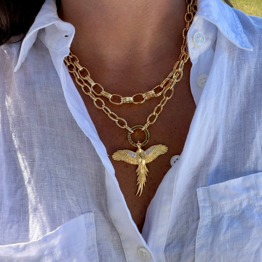 A close-up of a person wearing a layered gold necklace with a bird pendant, styled with a white linen button-up shirt.