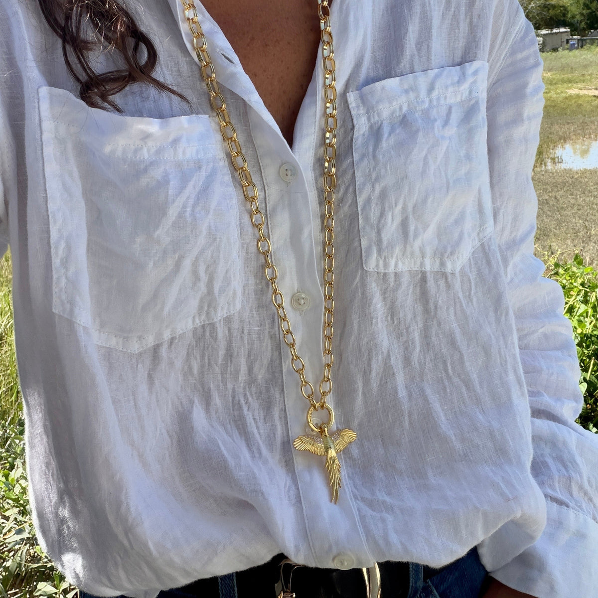 A person wearing a long gold chain necklace with a cross pendant, styled over a white button-up shirt in an outdoor setting.