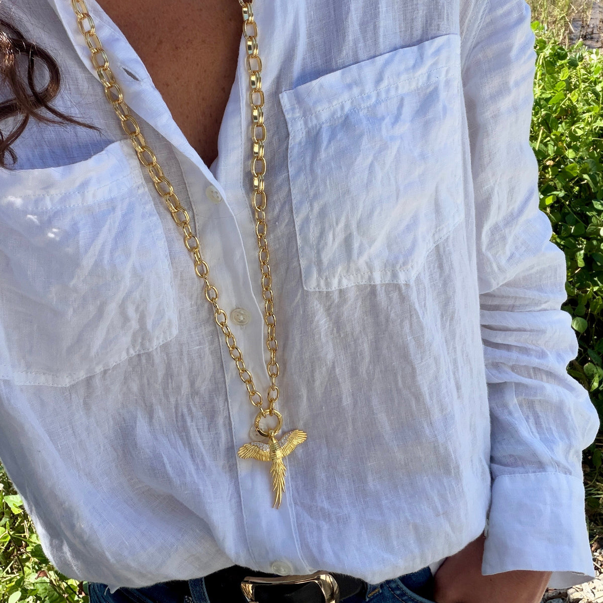 A close-up of a person wearing a white linen button-up shirt and a long gold chain necklace with a textured cross pendant, outdoors near greenery and water.